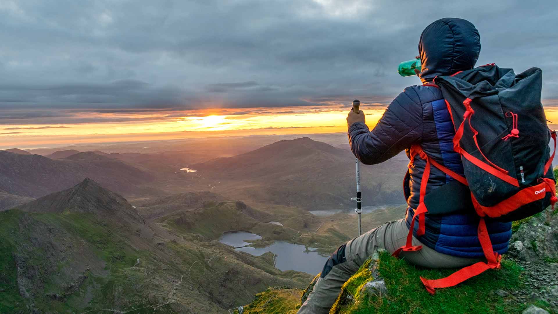 woman sitting on mountain in a heavy coat. She is watching the sunset in the distance.
